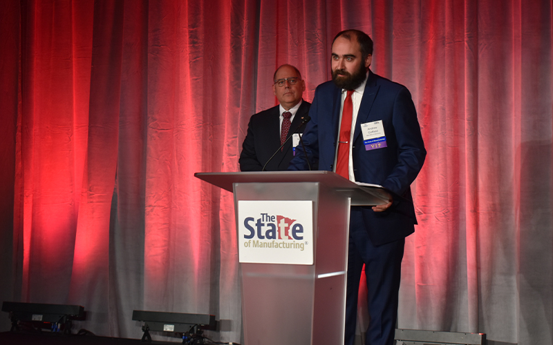 Andrew Coffield of the SBA's Office of Advocacy speaks at a podium bearing The State of Manufacturing® logo at Enterprise Minnesota's 2025 annual event, with a colleague standing behind him on stage.