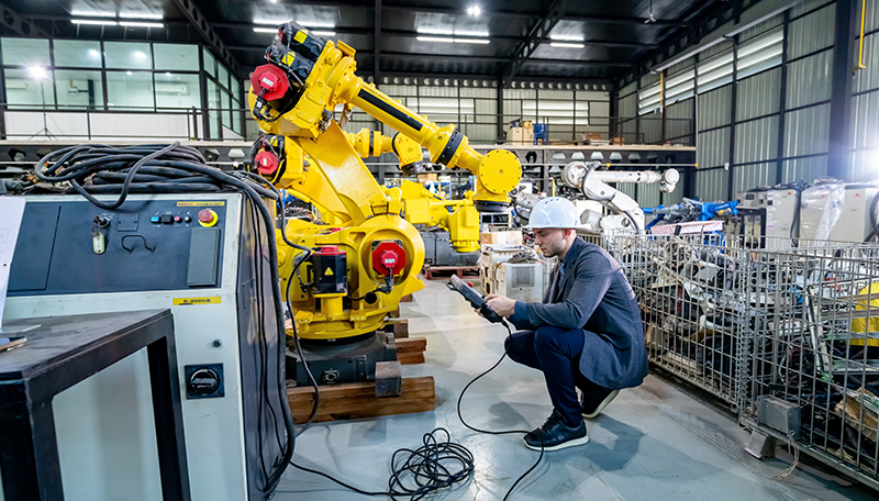 A engineer wearing a white hard hat crouches on a manufacturing floor, closely inspecting a large yellow industrial robotic arm while holding a control device.