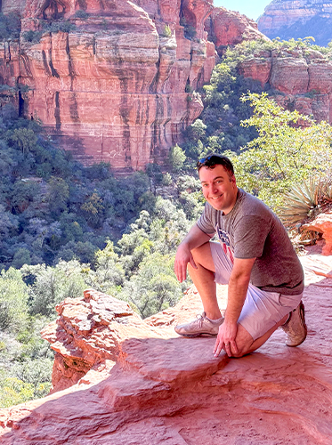 A smiling man crouches on a red rock ledge along a scenic hiking trail, with towering red rock formations and a lush green canyon valley visible in the background.