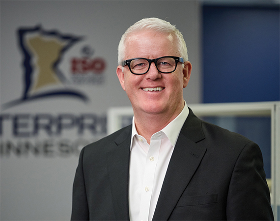 Headshot of a smiling man with white hair and black glasses wearing a dark blazer and white dress shirt, standing in front of an Enterprise Minnesota branded backdrop.