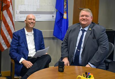 Enterprise Minnesota lobbyist Marty Seifert and Senator Rich Draheim seated together at a table in a legislative office, smiling with an American flag and Minnesota state flag visible in the background.