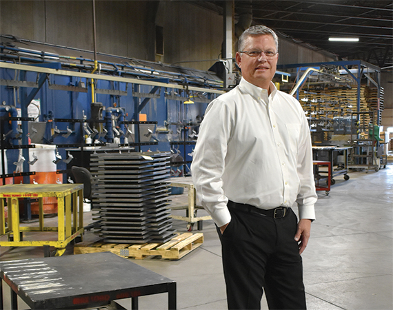 Gauthier Industries owner Mike Jensen stands on the floor of the company's manufacturing facility, with stacked metal components, industrial equipment, and a painting line visible in the background.