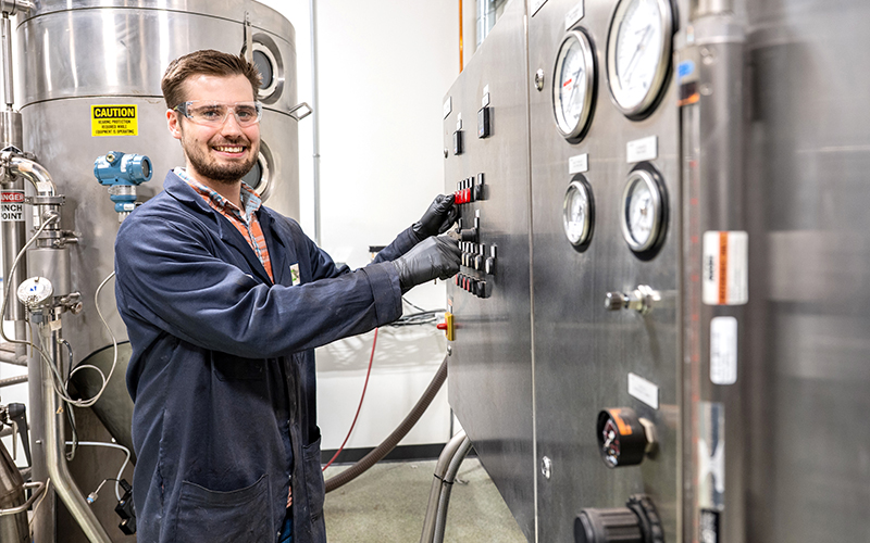 A smiling Niron Magnetics technician wearing a lab coat, safety glasses, and black gloves operates a stainless steel control panel next to industrial manufacturing equipment.