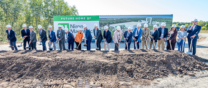 A large group of officials and dignitaries in white hard hats break ground with shovels in front of a Niron Magnetics sign reading "Future Home of Niron Magnetics" in Sartell, Minnesota.