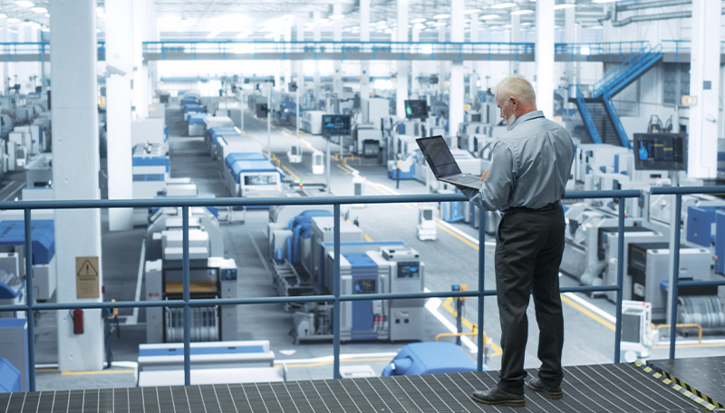 A senior business leader holding a laptop stands on an elevated mezzanine overlooking a vast, brightly lit modern manufacturing facility filled with rows of automated equipment.