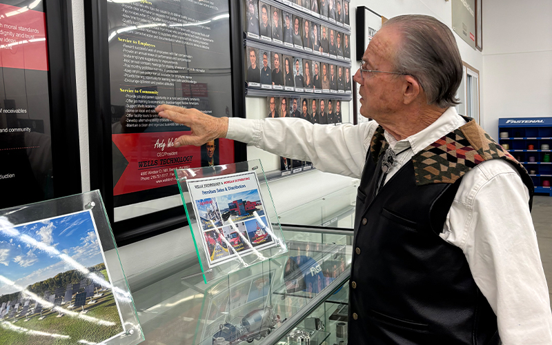 Wells Technology founder and CEO Andy Wells Sr. points to a large framed mission and values poster on the wall of the company's Bemidji facility, with employee photos and awards displayed nearby.