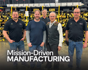 Five Wells Technology team members, including founder and CEO Andy Wells Sr., pose together on the manufacturing floor surrounded by yellow-tagged metal stock racks.