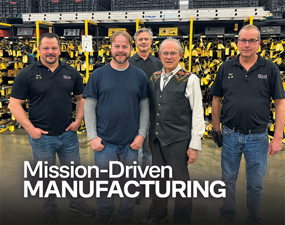 Five Wells Technology team members, including founder and CEO Andy Wells Sr., pose together on the manufacturing floor surrounded by yellow-tagged metal stock racks.